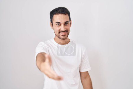 Foto de Hombre hispano guapo de pie sobre fondo blanco sonriendo amistoso ofreciendo apretón de manos como saludo y bienvenida. negocios exitosos. - Imagen libre de derechos