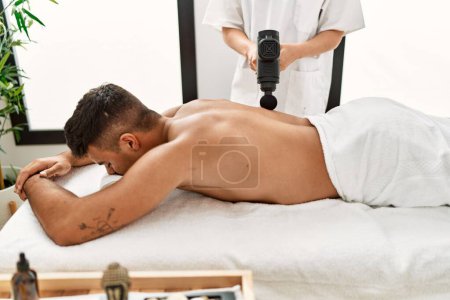 Foto de Joven hombre hispano relajado teniendo masaje de espalda usando pistola de percusión en el centro de belleza - Imagen libre de derechos