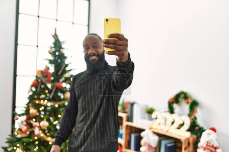 Téléchargez les photos : Jeune homme afro-américain faire selfie par le smartphone debout près de l'arbre de Noël à la maison - en image libre de droit