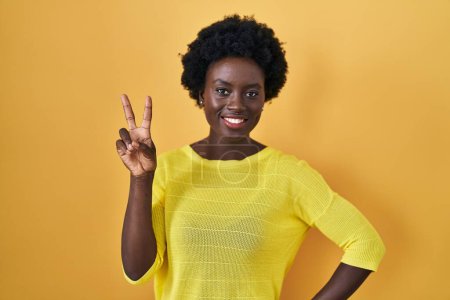 Téléchargez les photos : Jeune femme africaine debout sur le studio jaune souriant en regardant la caméra montrant les doigts faisant signe de victoire. numéro deux. - en image libre de droit