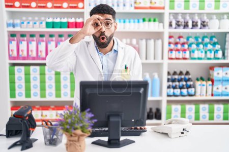 Photo for Hispanic man with beard working at pharmacy drugstore doing ok gesture shocked with surprised face, eye looking through fingers. unbelieving expression. - Royalty Free Image