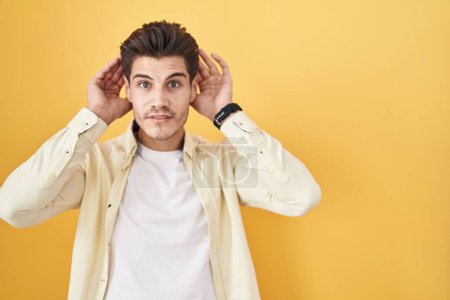 Photo for Young hispanic man standing over yellow background trying to hear both hands on ear gesture, curious for gossip. hearing problem, deaf - Royalty Free Image
