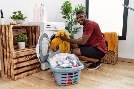 Téléchargez les photos : Jeune homme afro-américain nettoyage des vêtements à l'aide d'une machine à laver à la buanderie - en image libre de droit