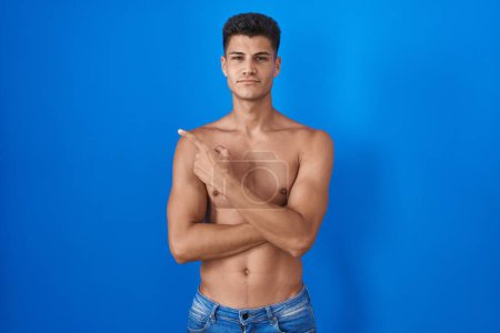 Photo for Young hispanic man standing shirtless over blue background pointing with hand finger to the side showing advertisement, serious and calm face - Royalty Free Image