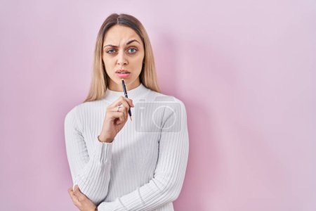 Photo for Young blonde woman standing holding pen over pink background skeptic and nervous, frowning upset because of problem. negative person. - Royalty Free Image