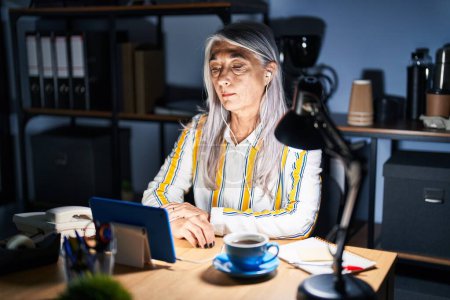Téléchargez les photos : Middle age woman with grey hair working at the office at night relaxed with serious expression on face. simple and natural looking at the camera. - en image libre de droit