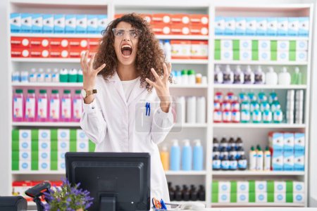 Téléchargez les photos : Hispanic woman with curly hair working at pharmacy drugstore crazy and mad shouting and yelling with aggressive expression and arms raised. frustration concept. - en image libre de droit