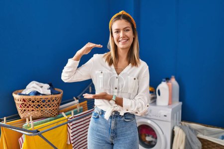 Téléchargez les photos : Young blonde woman at laundry room gesturing with hands showing big and large size sign, measure symbol. smiling looking at the camera. measuring concept. - en image libre de droit