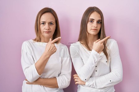 Téléchargez les photos : Middle age mother and young daughter standing over pink background pointing with hand finger to the side showing advertisement, serious and calm face - en image libre de droit