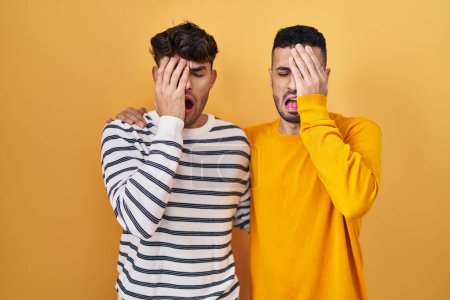 Foto de Young hispanic gay couple standing over yellow background yawning tired covering half face, eye and mouth with hand. face hurts in pain. - Imagen libre de derechos