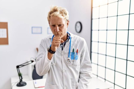 Foto de Young blond man wearing doctor uniform and stethoscope at clinic feeling unwell and coughing as symptom for cold or bronchitis. health care concept. - Imagen libre de derechos
