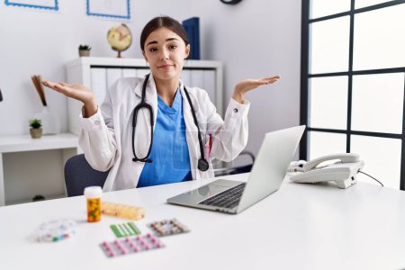 Photo for Young hispanic doctor woman wearing doctor uniform working at the clinic clueless and confused expression with arms and hands raised. doubt concept. - Royalty Free Image