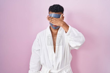 Photo for Young hispanic man wearing beauty face mask and bath robe covering eyes with hand, looking serious and sad. sightless, hiding and rejection concept - Royalty Free Image