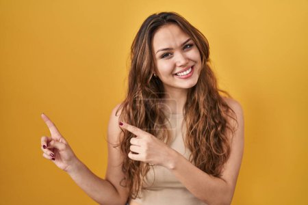 Foto de Young hispanic woman standing over yellow background smiling and looking at the camera pointing with two hands and fingers to the side. - Imagen libre de derechos