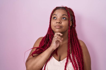 Téléchargez les photos : Femme afro-américaine avec des cheveux tressés debout sur fond rose pensant concentré sur le doute avec le doigt sur le menton et regardant vers le haut se demandant - en image libre de droit