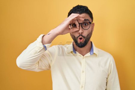 Foto de Hispanic young man wearing business clothes and glasses doing ok gesture shocked with surprised face, eye looking through fingers. unbelieving expression. - Imagen libre de derechos