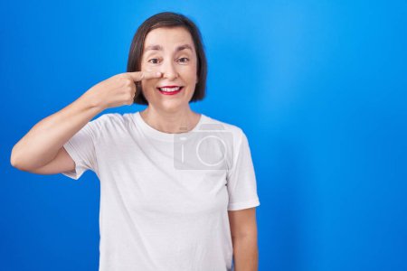 Téléchargez les photos : Middle age hispanic woman standing over blue background pointing with hand finger to face and nose, smiling cheerful. beauty concept - en image libre de droit
