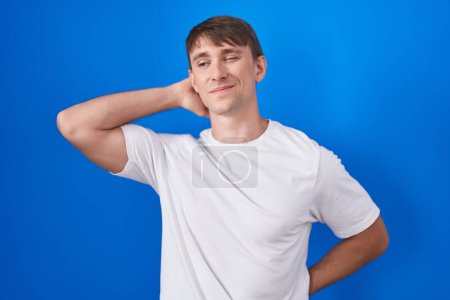 Photo for Caucasian blond man standing over blue background suffering of neck ache injury, touching neck with hand, muscular pain - Royalty Free Image