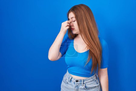 Foto de Redhead woman standing over blue background tired rubbing nose and eyes feeling fatigue and headache. stress and frustration concept. - Imagen libre de derechos