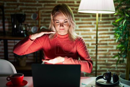 Téléchargez les photos : Blonde woman using laptop at night at home gesturing with hands showing big and large size sign, measure symbol. smiling looking at the camera. measuring concept. - en image libre de droit