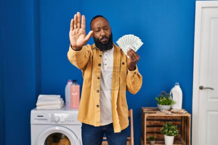 Téléchargez les photos : African american man holding money at laundry room with open hand doing stop sign with serious and confident expression, defense gesture - en image libre de droit