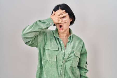 Téléchargez les photos : Jeune femme asiatique debout sur fond blanc regardant en état de choc couvrant le visage et les yeux avec la main, regardant à travers les doigts avec une expression embarrassée. - en image libre de droit
