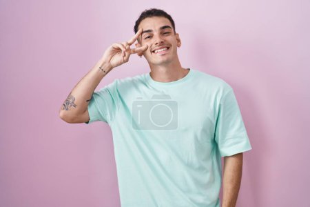Téléchargez les photos : Bel homme hispanique debout sur fond rose faisant symbole de paix avec les doigts sur le visage, souriant joyeux montrant la victoire - en image libre de droit