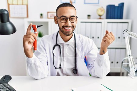 Photo for African american doctor man holding ear otoscope at the clinic screaming proud, celebrating victory and success very excited with raised arm - Royalty Free Image