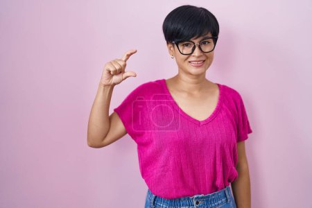 Téléchargez les photos : Young asian woman with short hair standing over pink background smiling and confident gesturing with hand doing small size sign with fingers looking and the camera. measure concept. - en image libre de droit