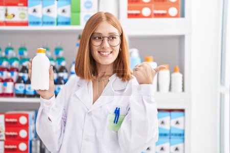 Photo for Young redhead woman working at pharmacy drugstore holding sun screen pointing thumb up to the side smiling happy with open mouth - Royalty Free Image