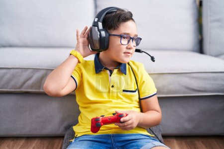 Téléchargez les photos : Jeune enfant hispanique jouant contrôleur de tenue de jeu vidéo portant des écouteurs souriant avec la main sur l'oreille à l'écoute d'une audience à la rumeur ou des commérages. concept de surdité. - en image libre de droit