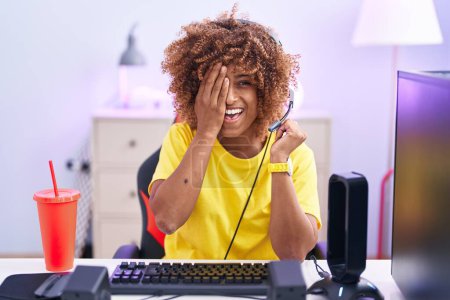 Téléchargez les photos : Young hispanic woman with curly hair playing video games wearing headphones covering one eye with hand, confident smile on face and surprise emotion. - en image libre de droit