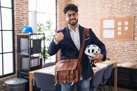 Foto de Hispanic man with beard holding bike helmet at the office smiling happy and positive, thumb up doing excellent and approval sign - Imagen libre de derechos