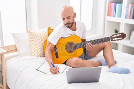 Téléchargez les photos : Jeune chauve compositeur de chanson jouant de la guitare classique assis sur le lit dans la chambre - en image libre de droit