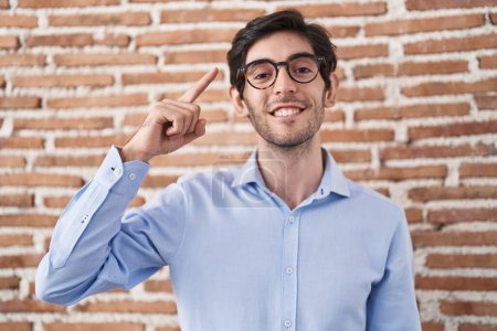 Téléchargez les photos : Young hispanic man standing over brick wall background smiling pointing to head with one finger, great idea or thought, good memory - en image libre de droit