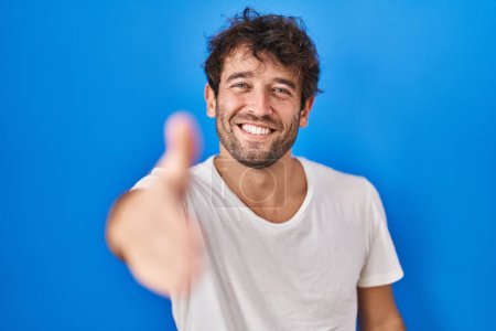 Foto de Joven hispano de pie sobre fondo azul sonriente amistoso ofreciendo apretón de manos como saludo y bienvenida. negocios exitosos. - Imagen libre de derechos
