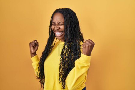 Photo for African woman standing over yellow background very happy and excited doing winner gesture with arms raised, smiling and screaming for success. celebration concept. - Royalty Free Image