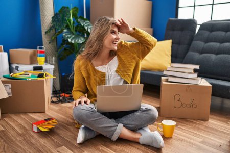 Foto de Young woman sitting on the floor at new home using laptop very happy and smiling looking far away with hand over head. searching concept. - Imagen libre de derechos