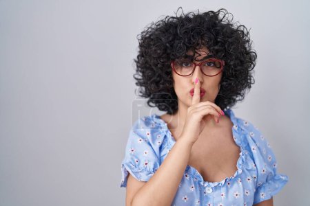 Photo for Young brunette woman with curly hair wearing glasses over isolated background asking to be quiet with finger on lips. silence and secret concept. - Royalty Free Image