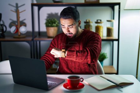 Téléchargez les photos : Jeune homme hispanique avec barbe utilisant ordinateur portable la nuit à la maison en regardant le temps de la montre inquiet, peur de se faire tard - en image libre de droit