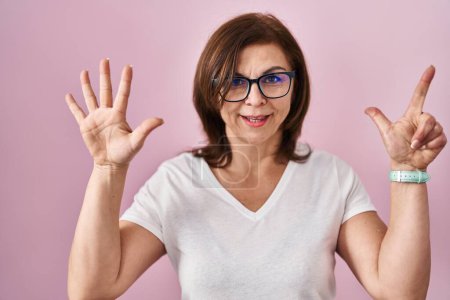 Foto de Mujer hispana de mediana edad de pie sobre fondo rosa mostrando y señalando hacia arriba con los dedos número siete mientras sonríe confiado y feliz. - Imagen libre de derechos