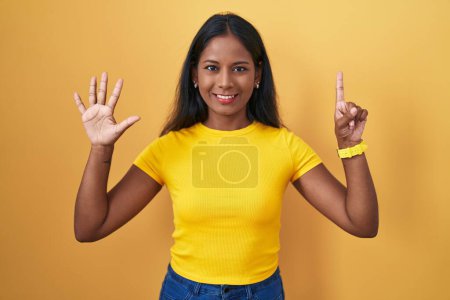 Foto de Mujer india joven de pie sobre fondo amarillo mostrando y señalando hacia arriba con los dedos número seis mientras sonríe confiado y feliz. - Imagen libre de derechos