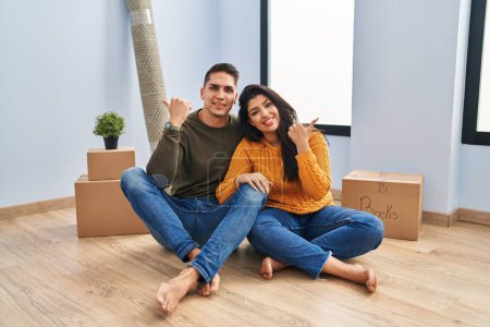 Foto de Young couple sitting on the floor at new home pointing to the back behind with hand and thumbs up, smiling confident - Imagen libre de derechos