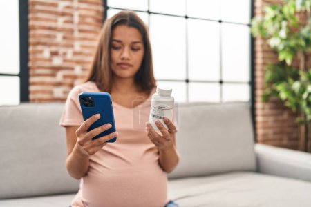 Téléchargez les photos : Jeune femme latine enceinte à l'aide d'un smartphone tenant bouteille de pilules à la maison - en image libre de droit