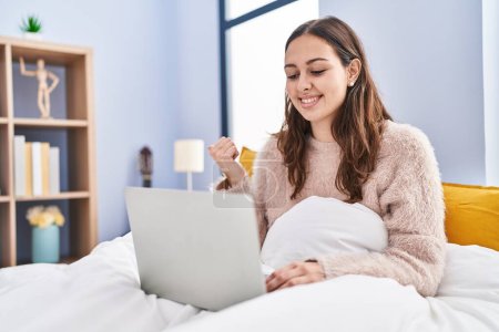 Téléchargez les photos : Jeune femme hispanique utilisant un ordinateur portable sur le lit pointant le pouce vers le côté souriant heureux avec la bouche ouverte - en image libre de droit