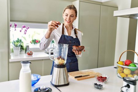 Téléchargez les photos : Jeune femme blonde souriante confiante versant des dates sur mélangeur à la cuisine - en image libre de droit