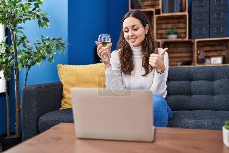 Photo for Young hispanic woman doing video call drinking white wine smiling happy and positive, thumb up doing excellent and approval sign - Royalty Free Image