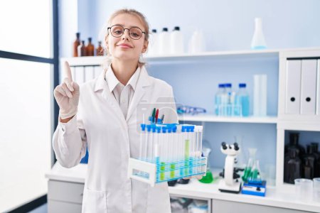 Photo for Young caucasian woman working at scientist laboratory holding samples smiling with an idea or question pointing finger with happy face, number one - Royalty Free Image