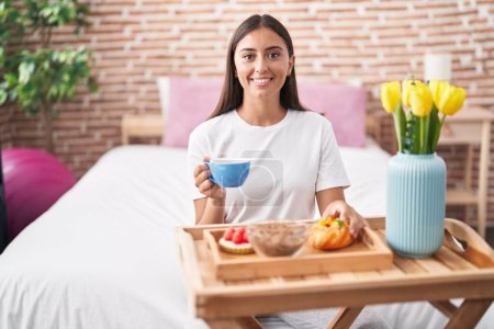 Téléchargez les photos : Young hispanic woman eating pastries for breakfast sitting on the bed smiling with a happy and cool smile on face. showing teeth. - en image libre de droit