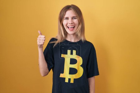 Téléchargez les photos : Blonde caucasian woman wearing bitcoin t shirt doing happy thumbs up gesture with hand. approving expression looking at the camera showing success. - en image libre de droit